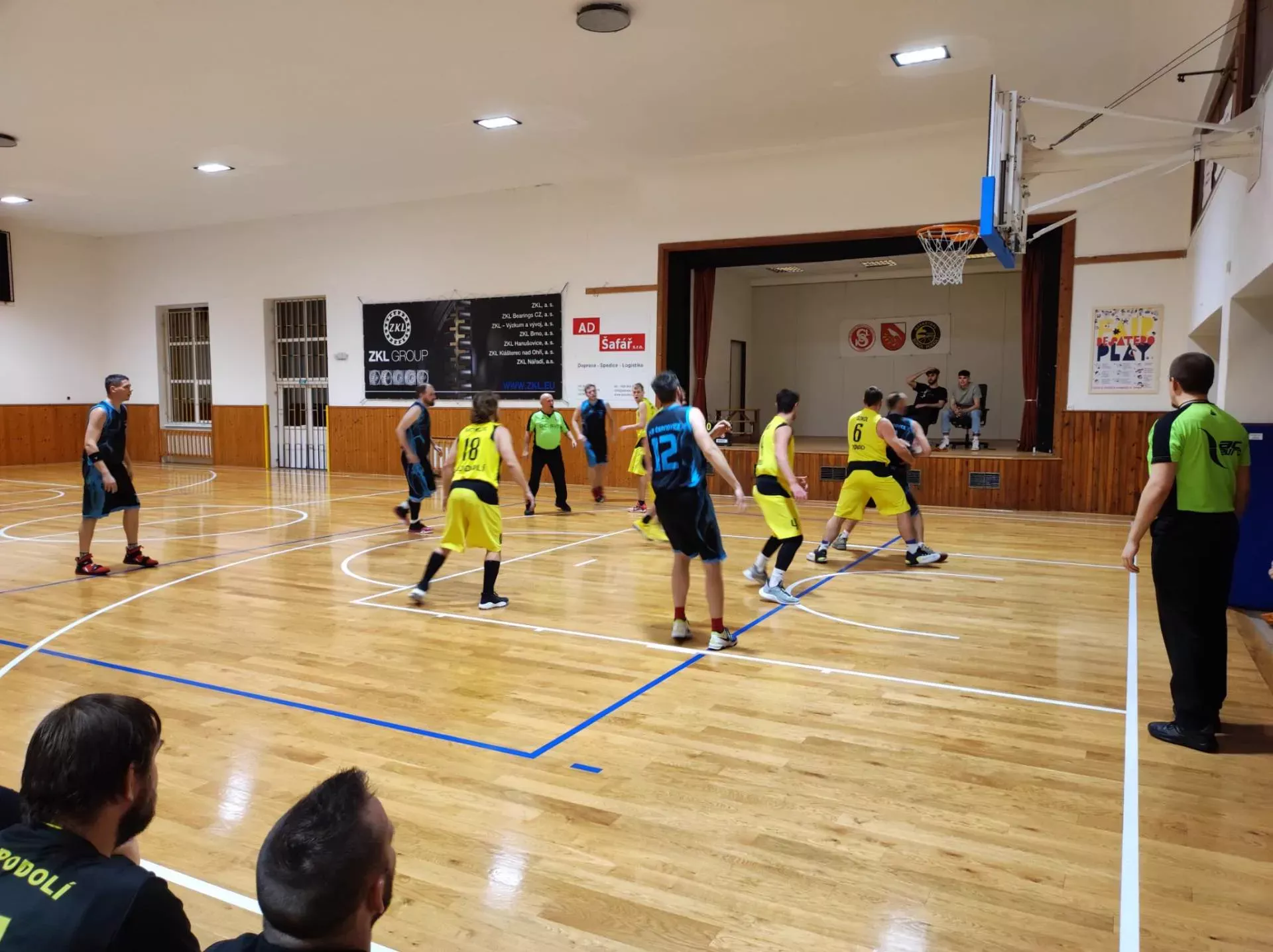 Muži B ve žlutých dresech v útoku proti soupeři v modrých dresech, v popředí lavička